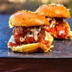 Two freshly grilled meatball sliders with tomato sauce and melted cheese served on a black plate outdoors.