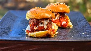 Two freshly grilled meatball sliders with tomato sauce and melted cheese served on a black plate outdoors.