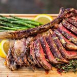 Sliced, perfectly cooked tomahawk steak served on a wooden cutting board with Z Grills logo, accompanied by fresh green asparagus and lemon slices, seasoned with herbs.