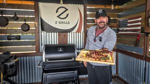 Happy chef in a Z Grills cap proudly holding a large, beautifully prepared Tomahawk steak in front of a Z Grills pellet grill, with an American flag and assorted cooking tools in the background.