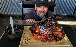 Cheerful chef presenting a glossy glazed ham on a wooden cutting board beside a Z Grills smoker, showcasing the results of expert grilling.