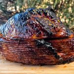 Glazed ham beautifully caramelized, presented on a wooden cutting board set against a natural outdoor backdrop.