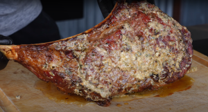 Close-up of a Tomahawk steak generously coated with a crust of herbs and garlic, resting on a cutting board after grilling.