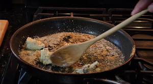 Stirring melted butter with herbs in a skillet over a gas stove, preparing a savory board sauce for a steak.