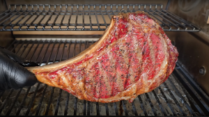 Placing a seasoned Tomahawk steak on the grill, showcasing the vibrant red meat with a generous coating of spices, handled by a chef wearing a black glove.