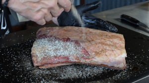 Chef liberally seasoning beef short ribs with a custom spice blend on a black cutting board.