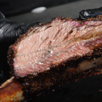 Close-up of a chef's gloved hand holding a perfectly cooked slice of beef short rib, showcasing the pink smoke ring and juicy interior.