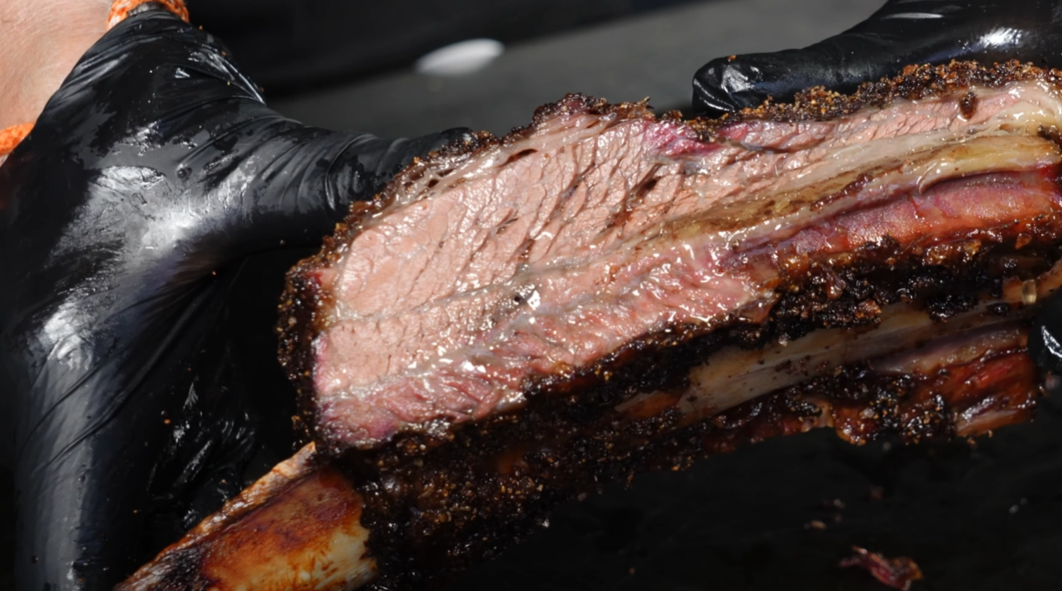 Close-up of a chef's gloved hand holding a perfectly cooked slice of beef short rib, showcasing the pink smoke ring and juicy interior.