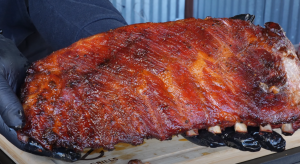 Grilled ribs with a glossy, caramelized crust being held with gloved hands over a Z Grills cutting board, showcasing the deep, rich barbecue color.