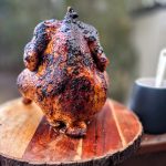 Perfectly roasted beer can chicken standing upright on a wooden board, with a crisp, golden-brown skin, cooked on a grill.