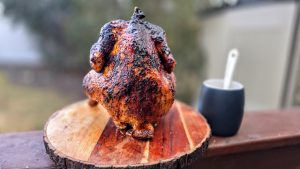 Perfectly roasted beer can chicken standing upright on a wooden board, with a crisp, golden-brown skin, cooked on a grill.