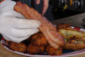 A gloved hand holds up a slice of freshly grilled candied bacon, showcasing its golden-brown sugar crust, with smoked sausage balls and loaded potato skins in the background.