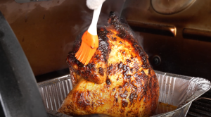 A close-up view of a golden-brown beer can chicken being basted with a bright orange basting brush, highlighting the juicy, crispy skin.