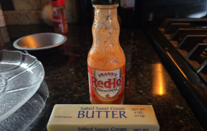 Bottle of Frank's RedHot sauce and a block of salted butter on a kitchen counter, essential ingredients for a spicy basting sauce.