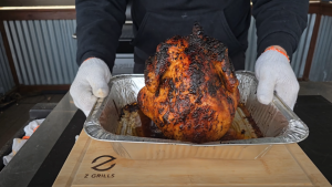 A perfectly roasted beer can chicken being lifted from a grill tray by a person wearing heat-resistant gloves.