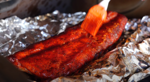 A brush applying a rich, glossy BBQ sauce on a perfectly cooked rib, highlighting the vibrant red color and juicy texture of the meat.