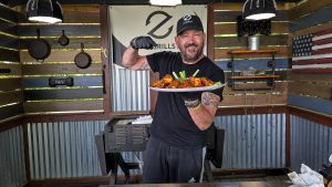 Man holding a plate of freshly grilled buffalo chicken wings in an outdoor kitchen featuring a Z Grills smoker.