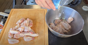 Raw chicken wings being coated with baking powder in a metal mixing bowl, with additional wings on a cutting board beside it.
