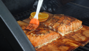 A brush applying honey orange glaze onto cedar plank salmon cooking on a grill.