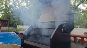 Z Grills pellet smoker with the lid open and smoke rising, set outdoors near a swimming pool.