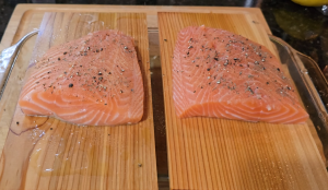 Two raw salmon fillets seasoned with cracked pepper and herbs, placed on soaked cedar planks.