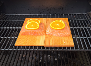 Raw salmon fillets topped with orange slices placed on cedar planks over a pellet grill.