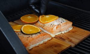 Raw salmon fillets placed on cedar planks with orange slices and seasoning, being arranged on a grill.