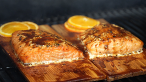 Two fillets of cedar plank salmon cooking on a grill, glazed and topped with orange slices.