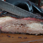 Juicy sliced brisket showing smoke ring, bark, and rendered fat layers, being cut with a slicing knife on a wooden board.