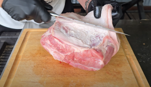 Person trimming and removing the fat cap from a raw pork shoulder on a wooden cutting board.