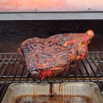 Smoked pork shoulder resting on a pellet grill with a drip pan catching juices below.