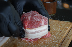 Bacon-wrapped filet mignon being seasoned with salt on a wooden cutting board