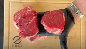 Two raw filet mignon steaks held by gloved hand over a Z Grills cutting board.