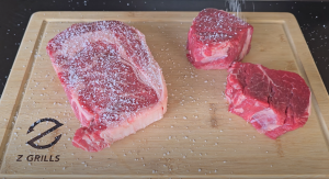 Ribeye and filet mignon steaks on a Z Grills cutting board being seasoned with kosher salt.