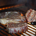 Gloved hand holding a seared filet mignon over flames on a Z Grills pellet grill, with ribeye and another filet searing on the grates.
