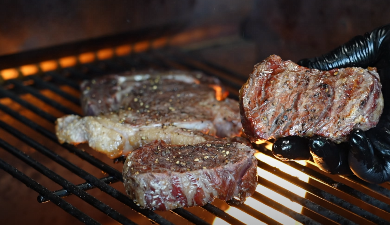 Gloved hand holding a seared filet mignon over flames on a Z Grills pellet grill, with ribeye and another filet searing on the grates.
