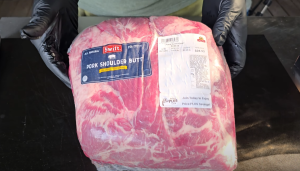 Gloved hands holding a packaged fresh pork shoulder butt on a black cutting board.