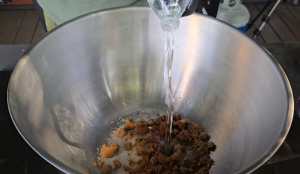 Water being poured into a metal mixing bowl with brown sugar, salt, and spices for pork brine.