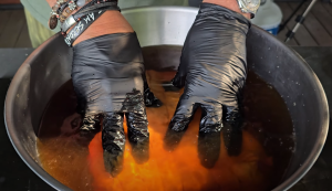 Hands in black gloves submerging pork butt into a seasoned brine in a large bowl.
