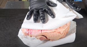 Gloved hands patting a pork butt dry with paper towels before seasoning.